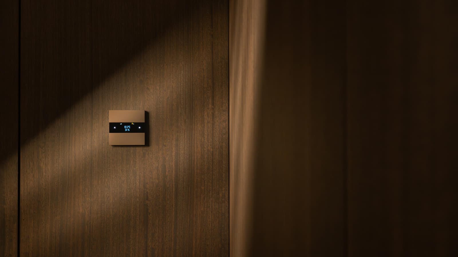 Basalte Deseo thermostat in bronze, flush-mounted on a walnut wall and showing 19.5 °C, lit by a shaft of daylight.
