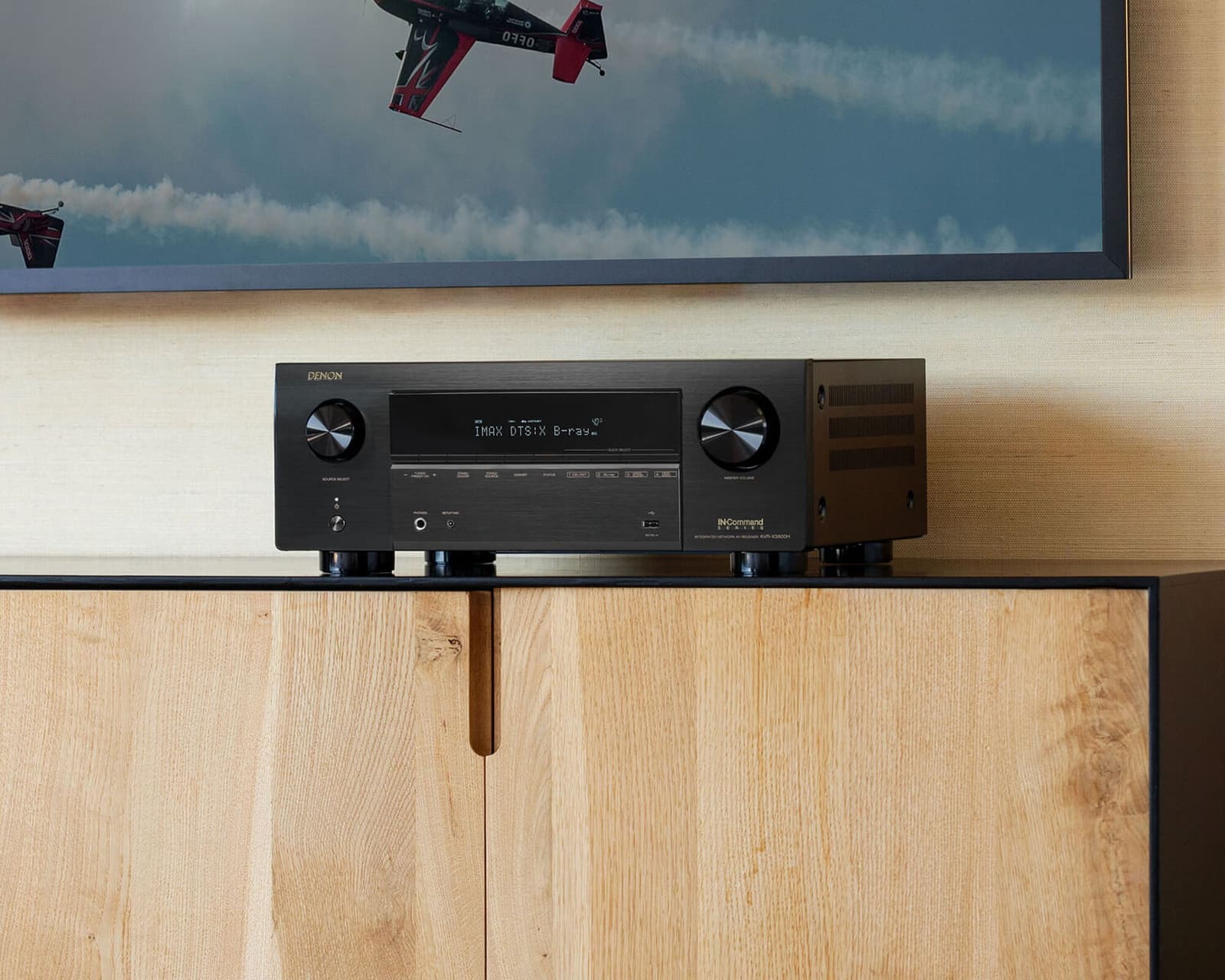 Close view of a Denon AVR-X3800H on an oak sideboard, its display reading IMAX DTS:X, below a wall-mounted TV.
