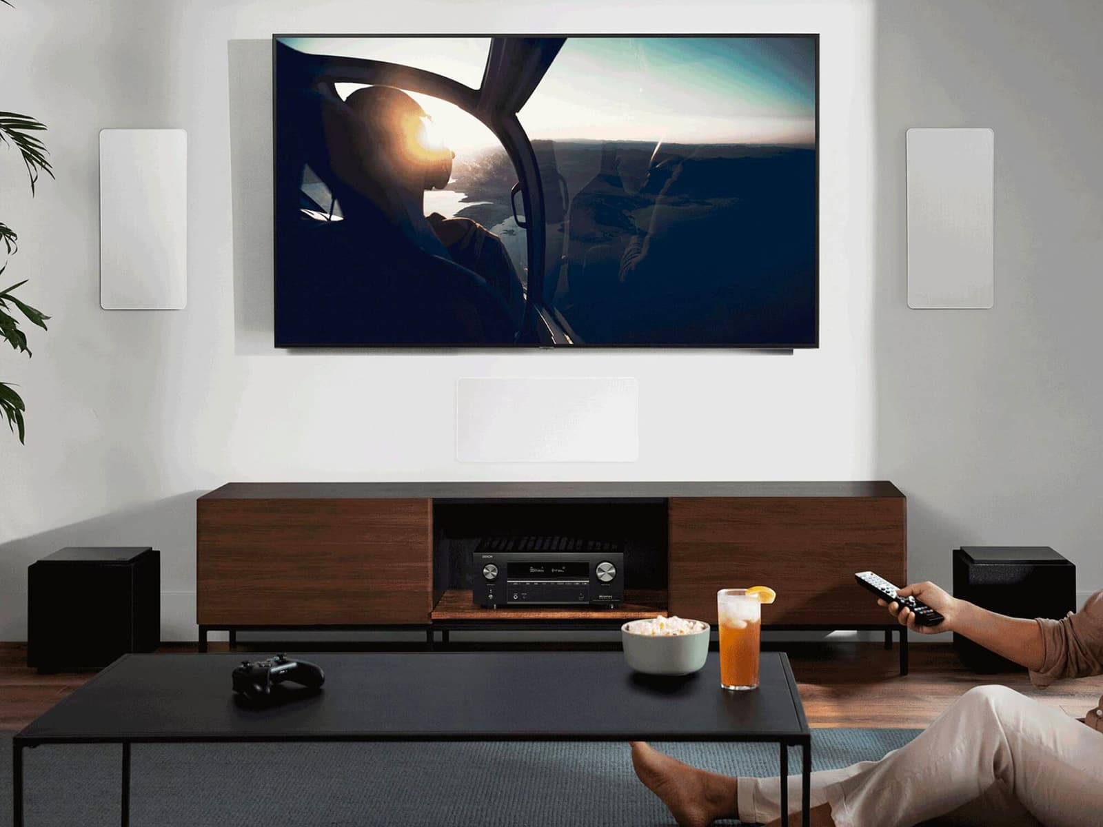Black Denon AVR-X3800H receiver in a walnut media console below a wall-mounted TV, with on-wall speakers and subwoofers in a living room.
