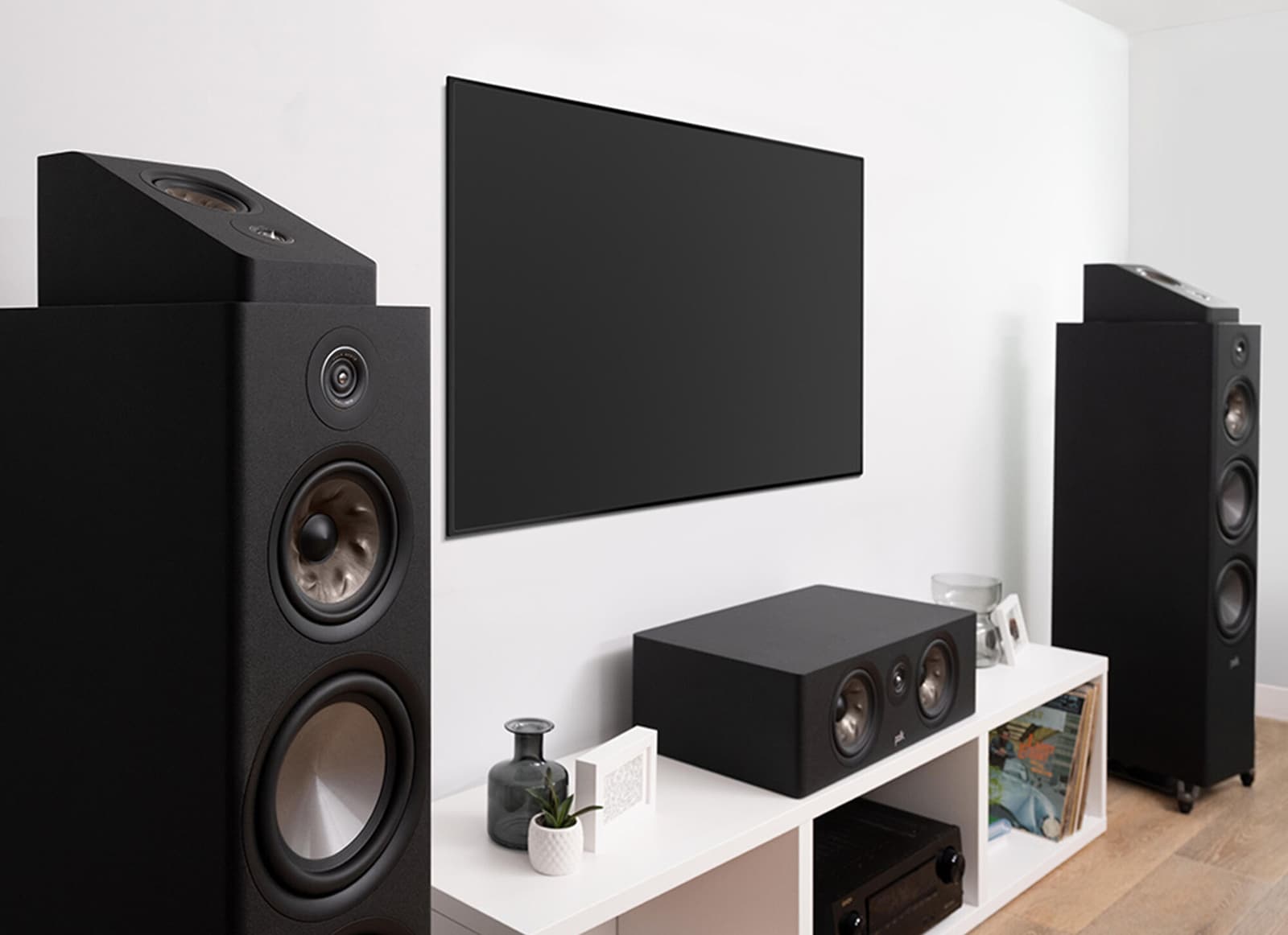 Two black Polk Reserve tower speakers and a Polk centre channel around a wall-mounted TV in a white family room.