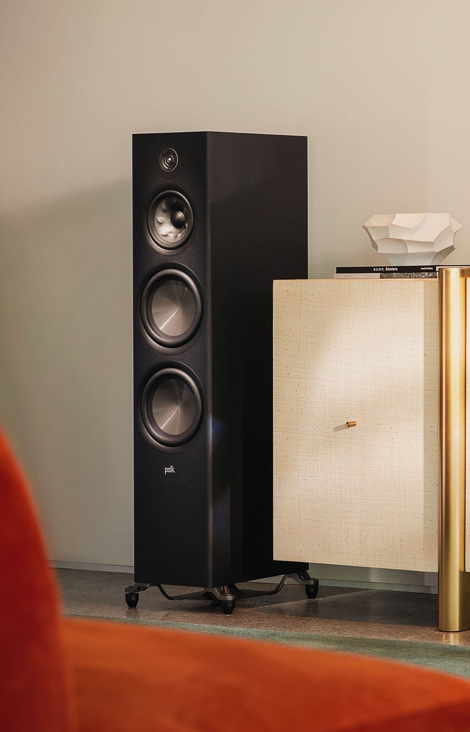 Black Polk Reserve tower speaker with its grille off, beside a raffia-fronted cabinet on a terrazzo floor.