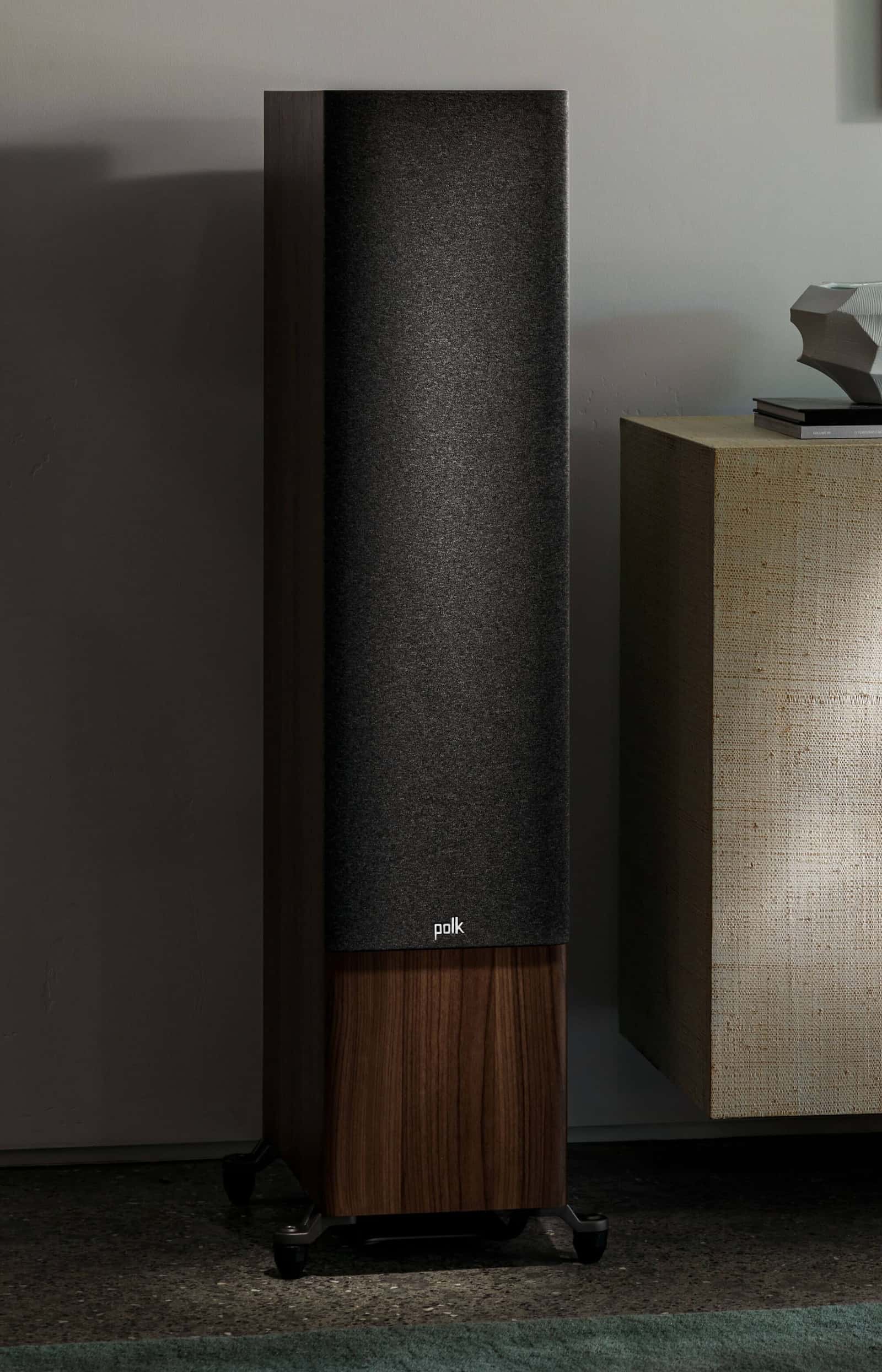 Polk Reserve tower speaker in walnut with grille fitted, against a taupe plaster wall beside a linen-fronted cabinet.
